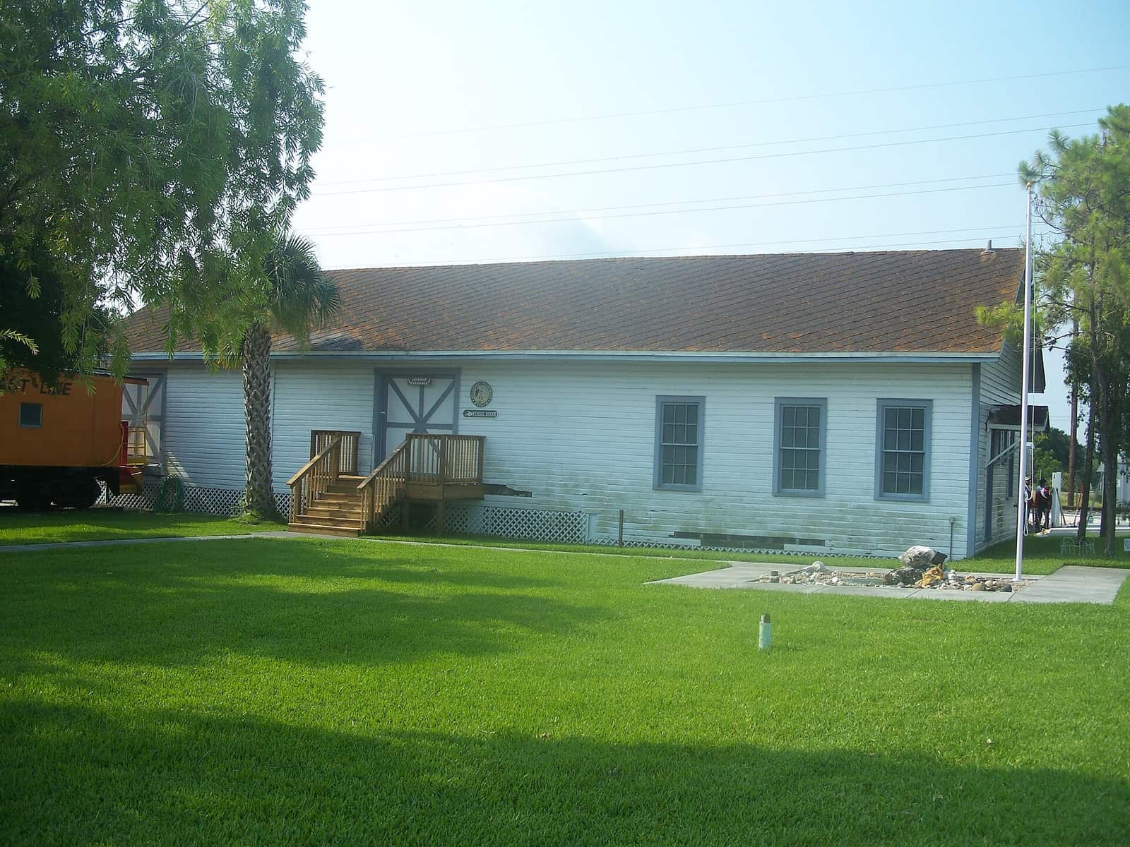 Light blue wooden museum building with wooden steps, a flagpole, a lawn and a caboose at left.