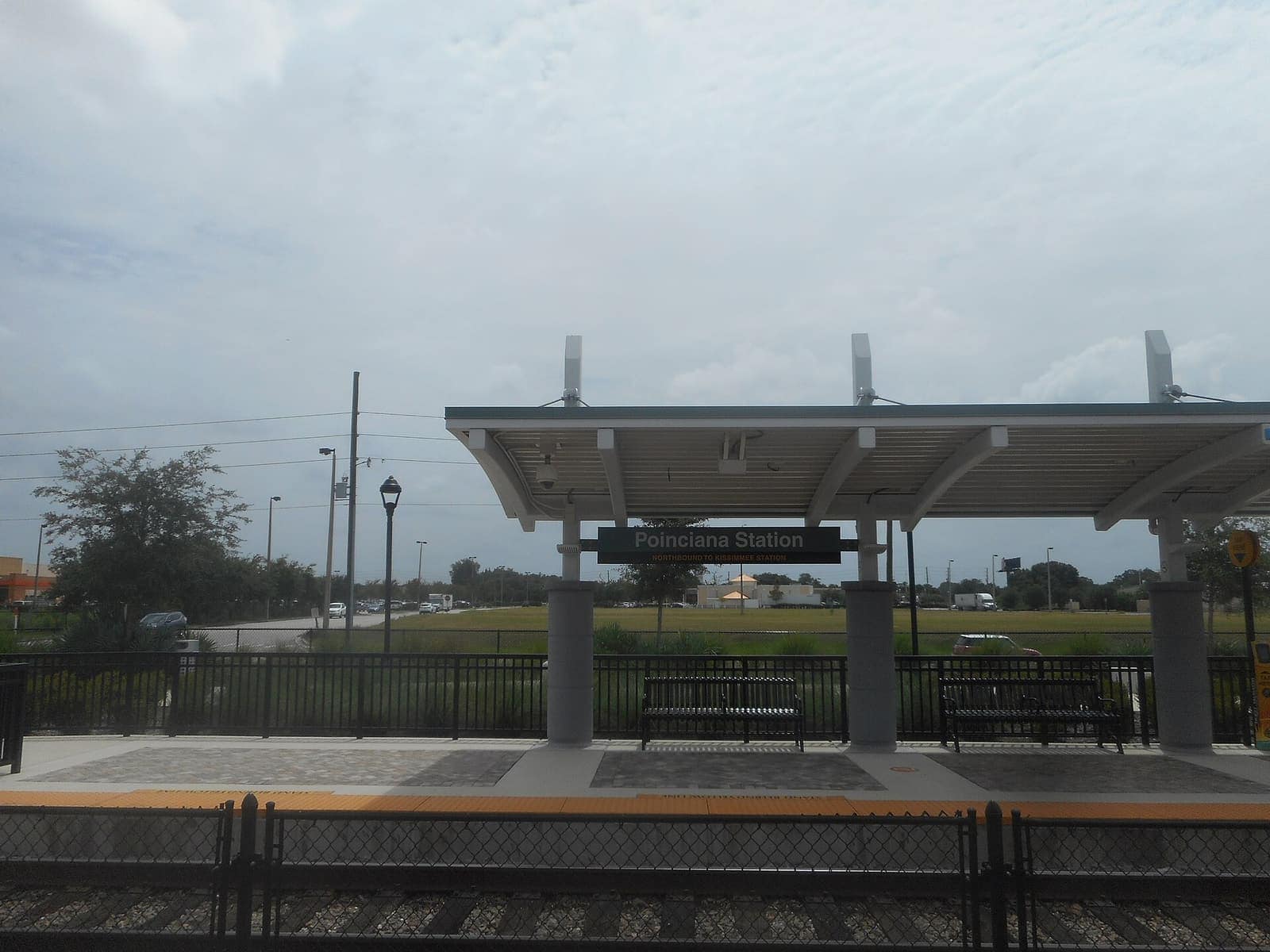 Covered railway platform with a Poinciana Station sign, benches, railing, and an open grassy field beyond.