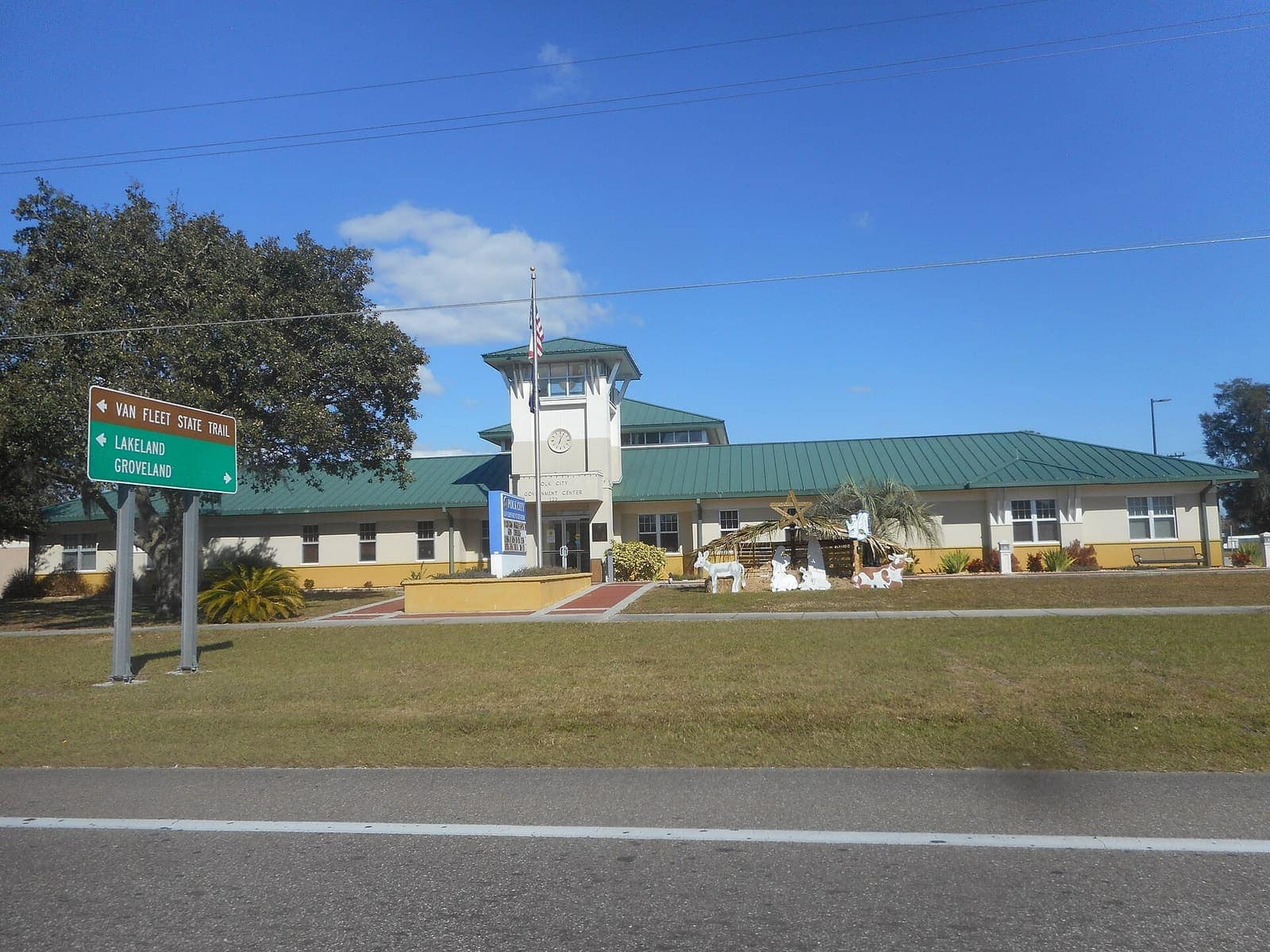 Single-storey government building with green metal roof, flagpole and a green road sign under a clear blue sky.