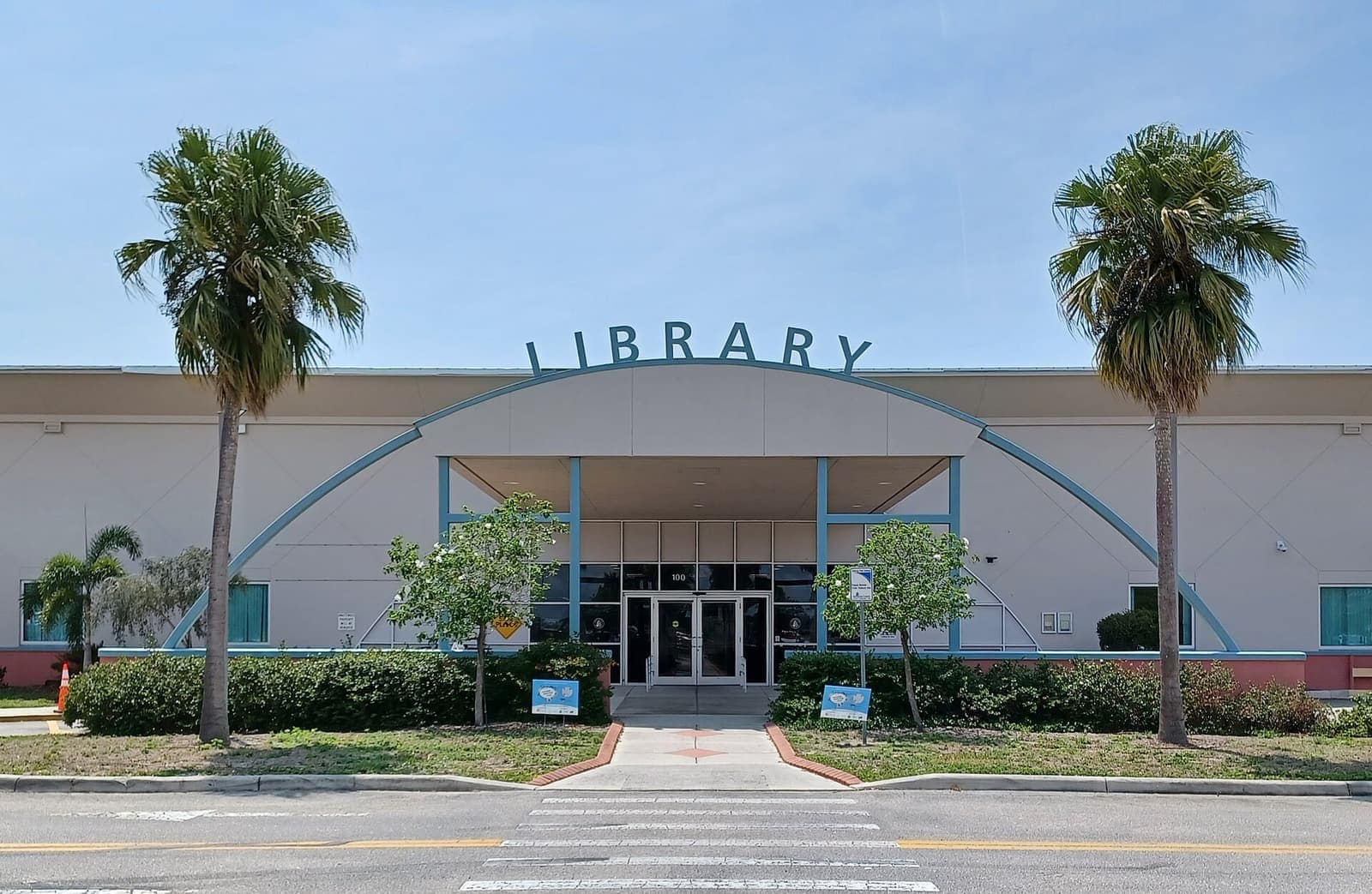 Front facade of a single-story library with palm trees and a sign reading LIBRARY under a blue sky.