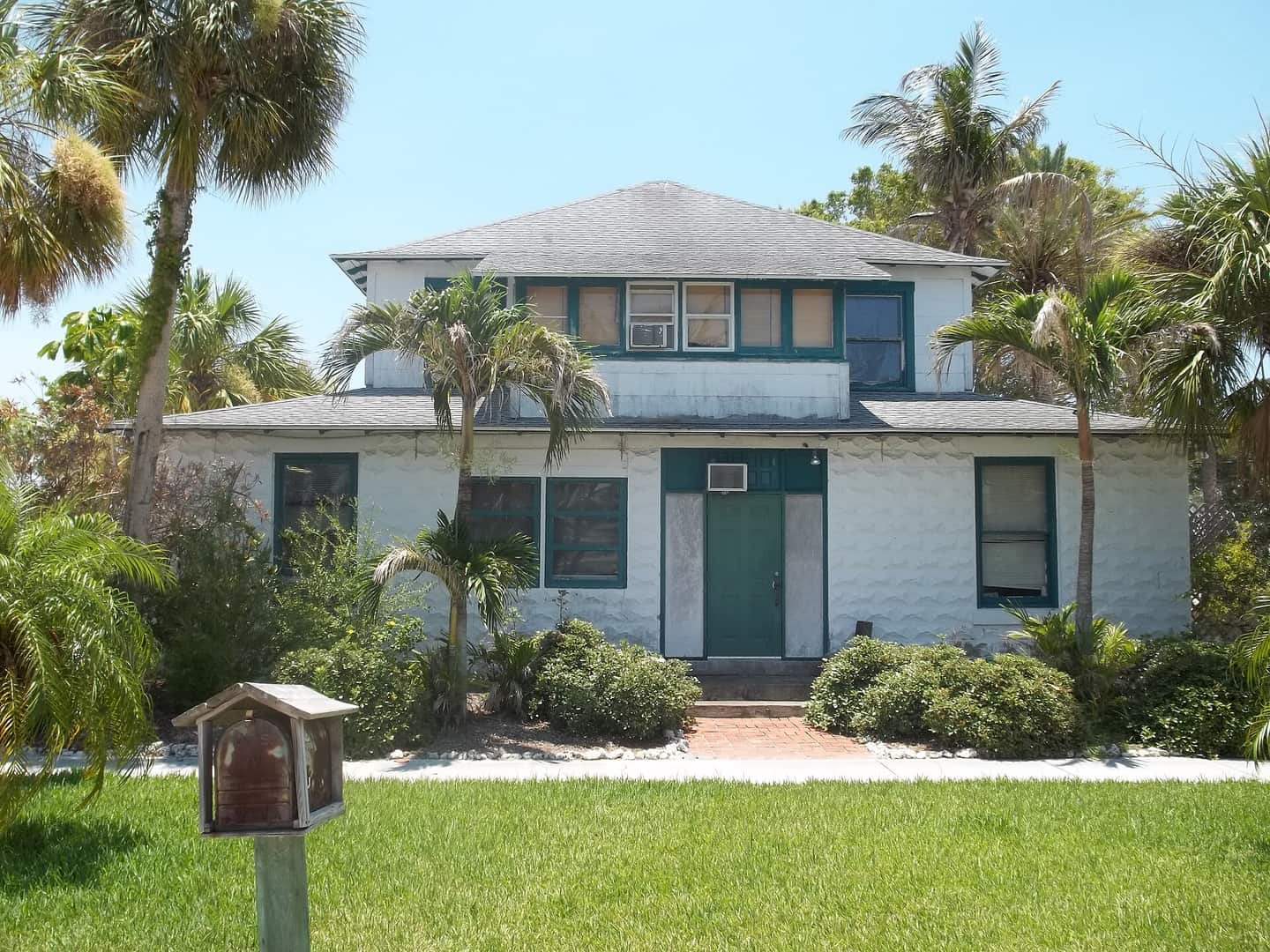 Two-storey white historic house with a green-trimmed entrance, palm trees, and a green lawn in daylight.