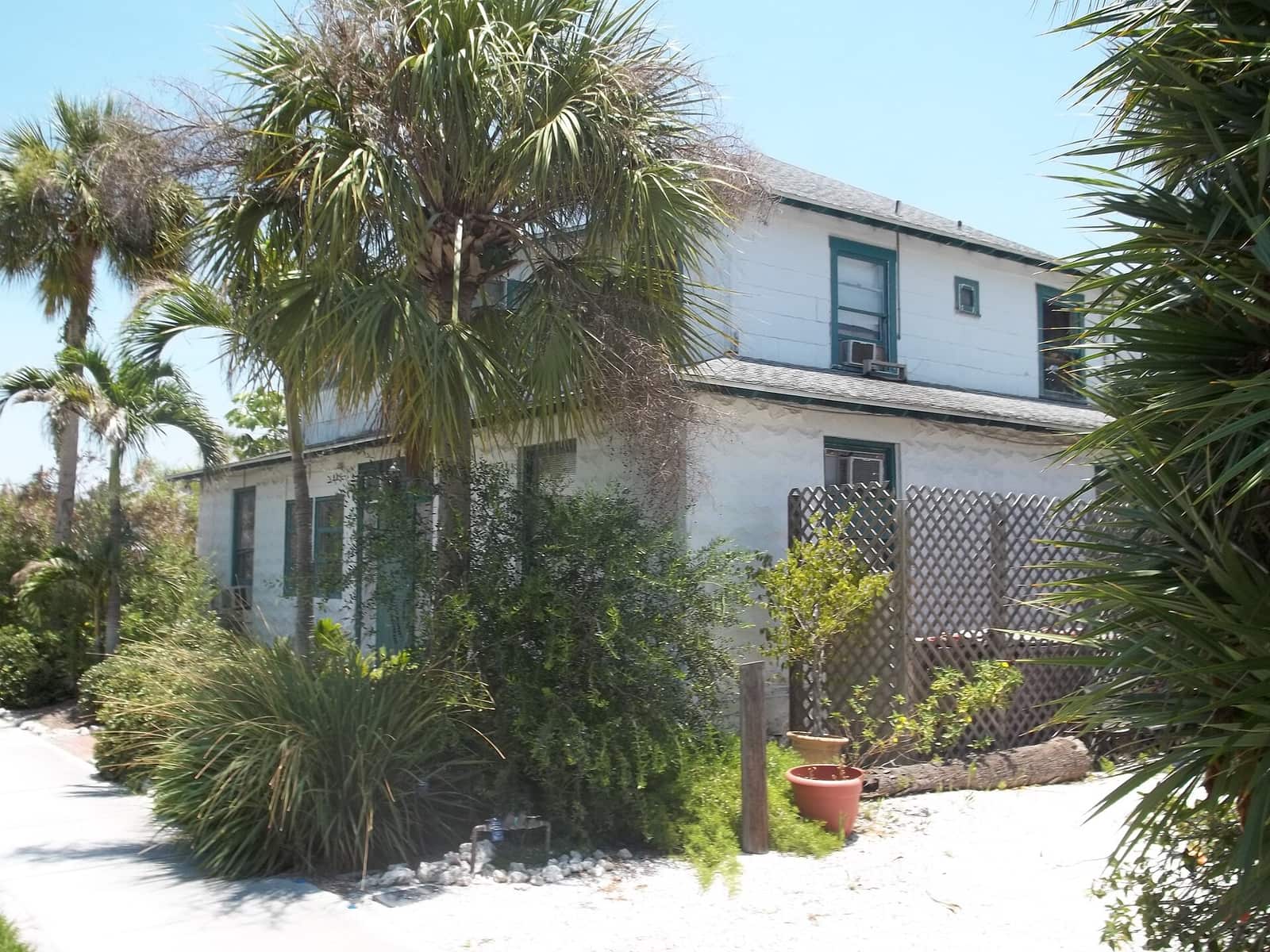 Two-storey white historic house surrounded by palms and shrubs along a sandy path in daylight.