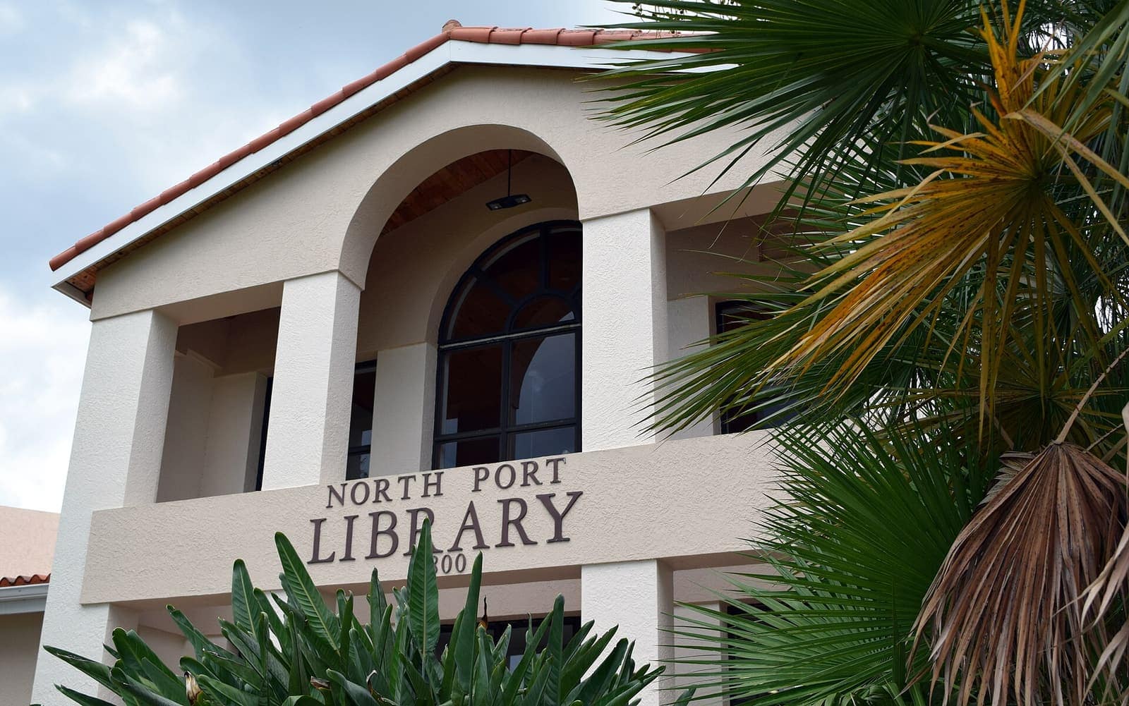 Cream stucco North Port Library building with arched windows, red tile roof and palm fronds beside it.