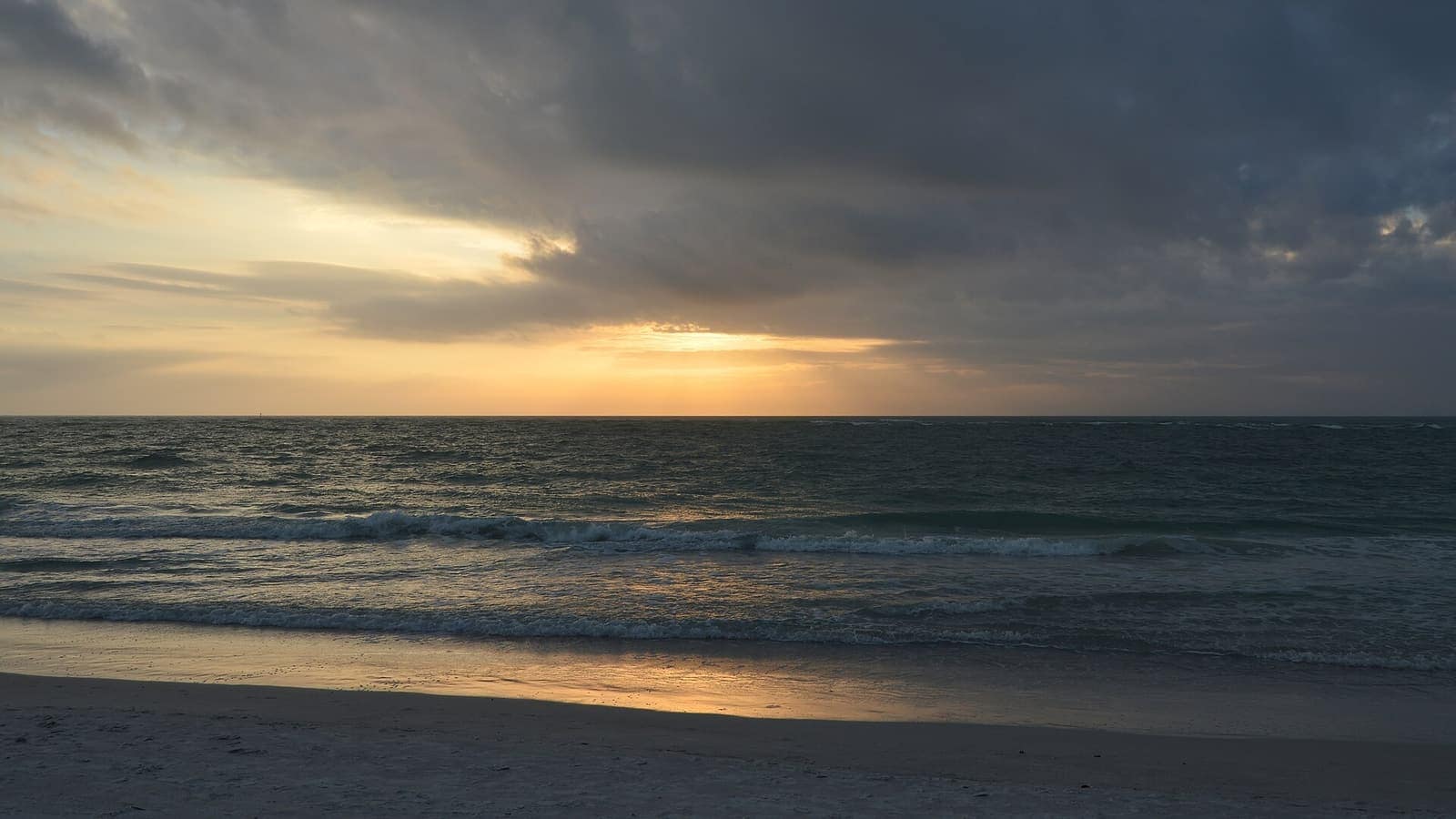 Gulf of Mexico waves at sunset under dark clouds, with a wet sand beach in the foreground.