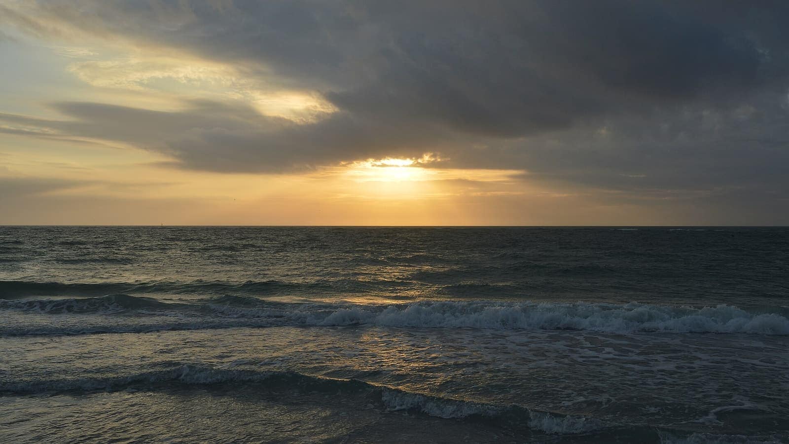 Sun breaking through clouds over the Gulf of Mexico at sunset, with small waves on the water.