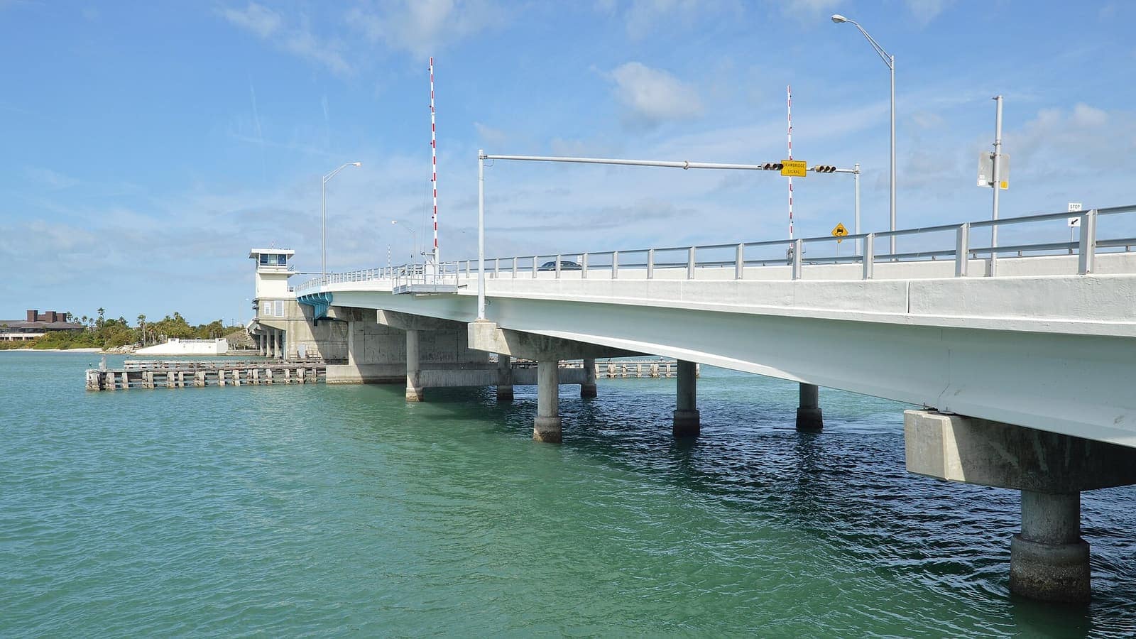 Concrete bridge with metal railings and a control tower stretches over turquoise water on a clear day.