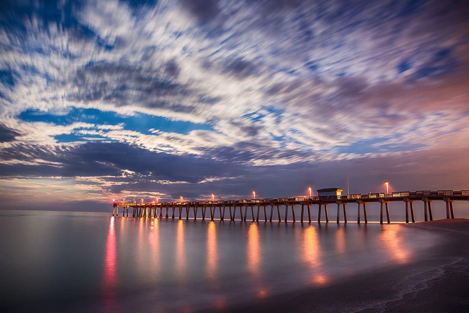 Long wooden fishing pier with lit posts extending over calm water under streaked clouds at dusk.