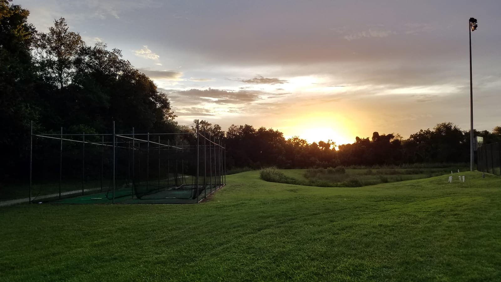 Golden sunset over an open green softball field with a batting cage and tall light pole.