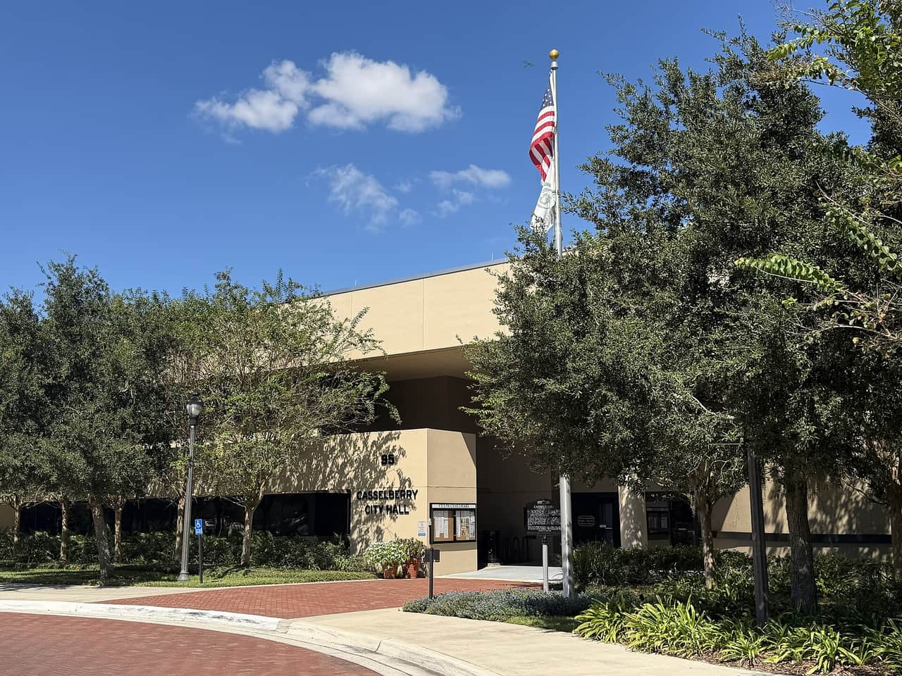 Tan city hall building with an American flag, leafy trees, and a brick walkway under blue sky.