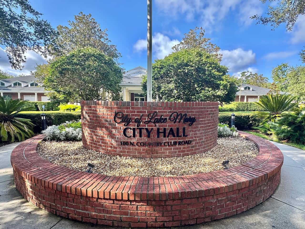 Curved brick city hall sign with dark lettering surrounded by shrubs, palms and a brick planter ring.