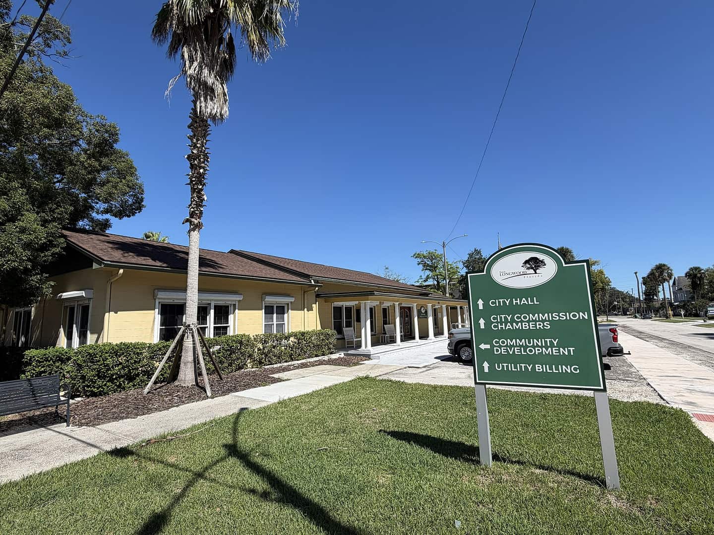 Yellow single-storey city hall building beside a green directional sign and a tall palm under blue sky.
