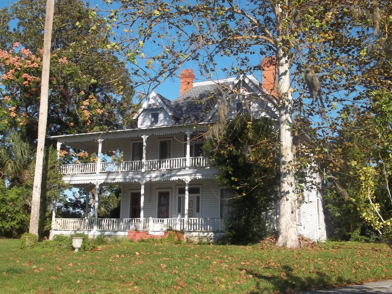 Two-story white historic house with wraparound porches, brick chimneys, and a large tree beside it.