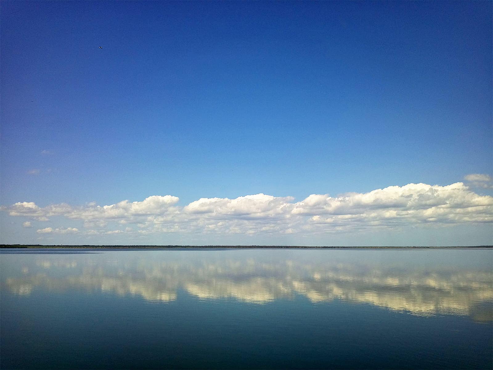 Calm lake reflecting a line of white clouds beneath a deep blue sky.