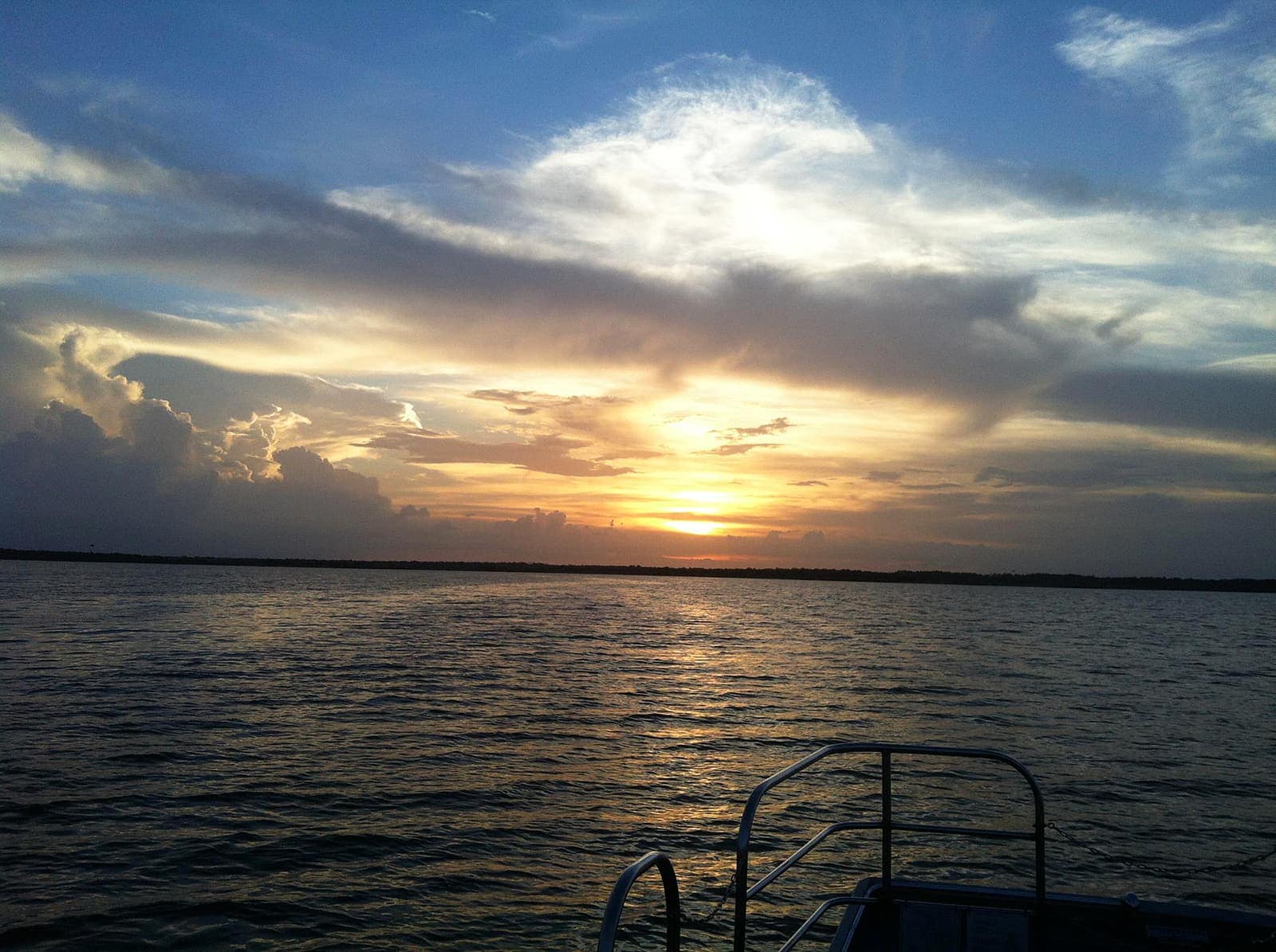 Orange sunset glowing through clouds over a calm lake, with a boat railing in the foreground.