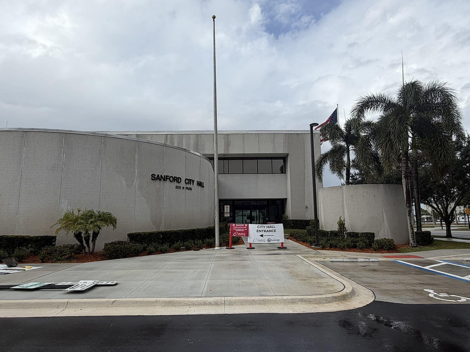 Modern concrete city hall building with a curved facade, palm trees, flagpole and entrance drive under cloudy sky.