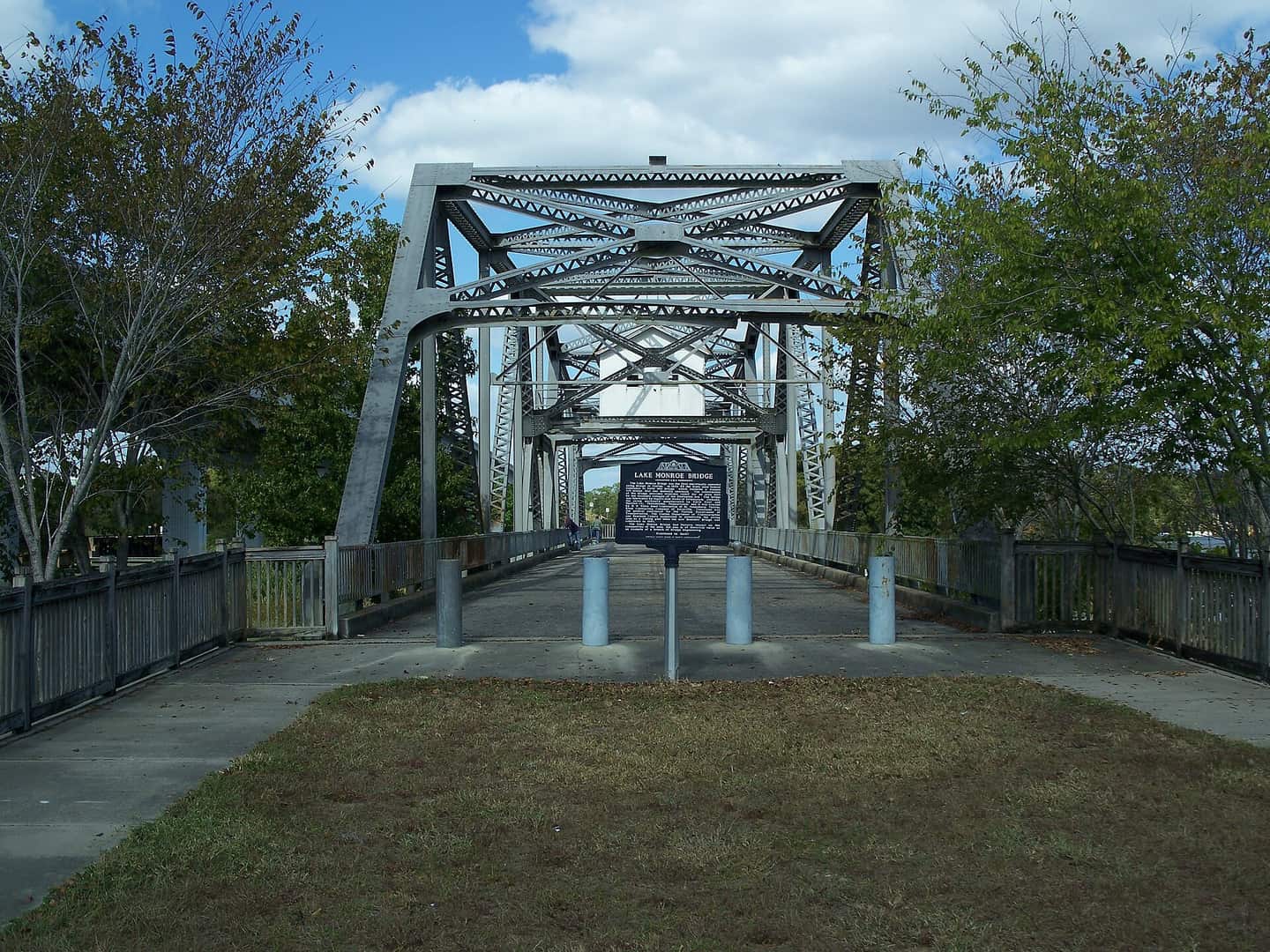 Historic steel truss bridge with a walkway and a historical marker sign at its center under blue sky.