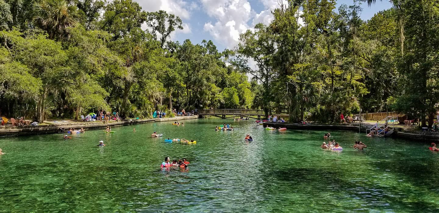 Clear green spring pool with people floating on tubes beneath shade trees and a footbridge.