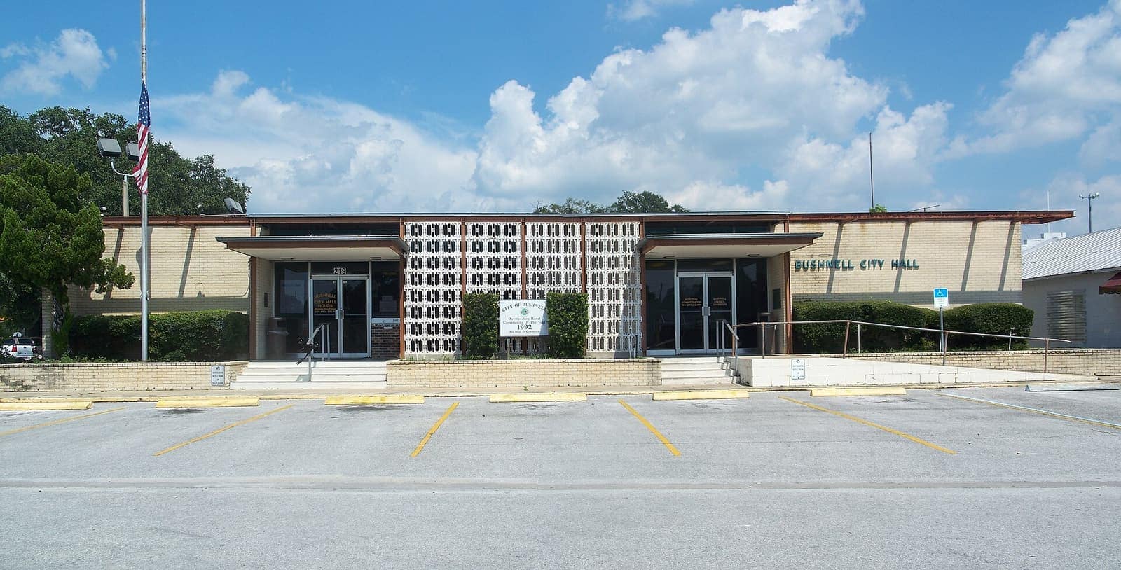 Single-storey tan city hall building with an American flag and a large empty parking lot.