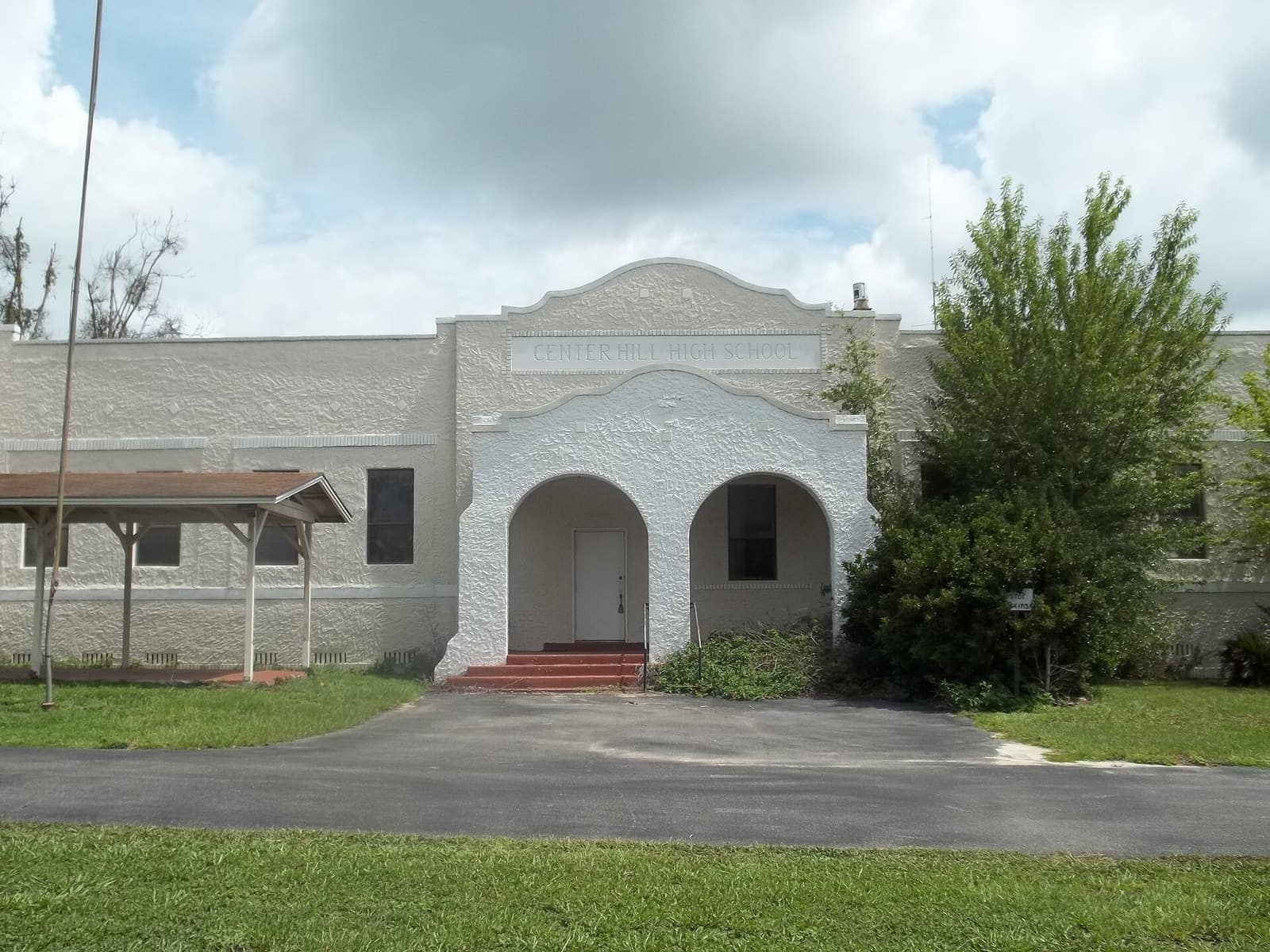 Whitewashed stucco school building with arched entrance and a paved driveway under cloudy sky.