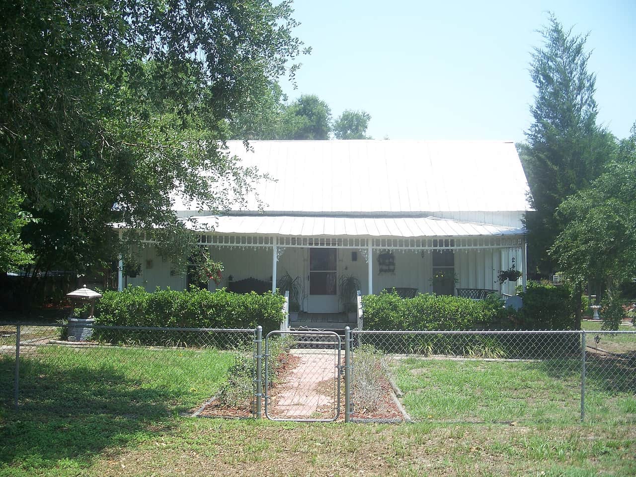 White-roofed frame cottage with a front porch behind a chain-link fence and green shrubs.