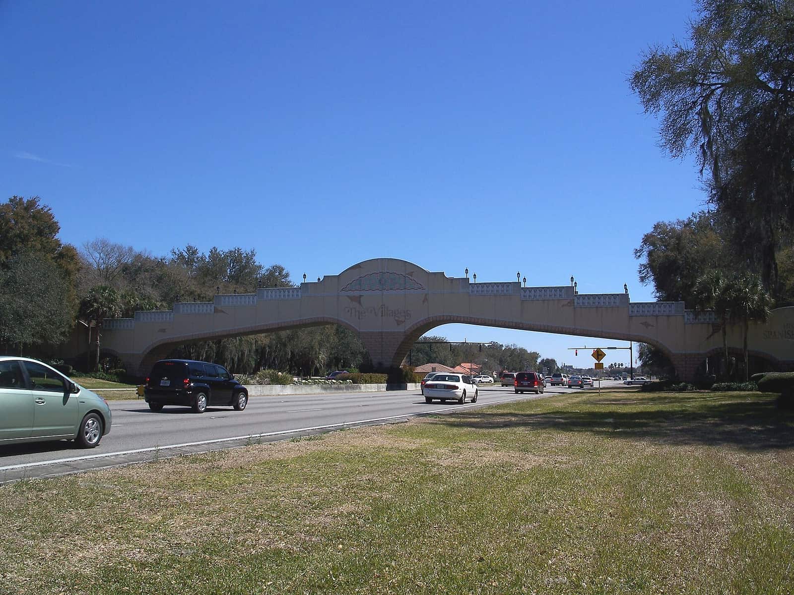 Tan concrete arched golf cart bridge spans a multi-lane road under a clear blue sky with cars passing.
