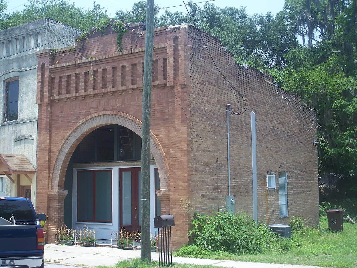 Small historic brick bank building with a tall arched entrance, shrubs and a parked dark vehicle at left.