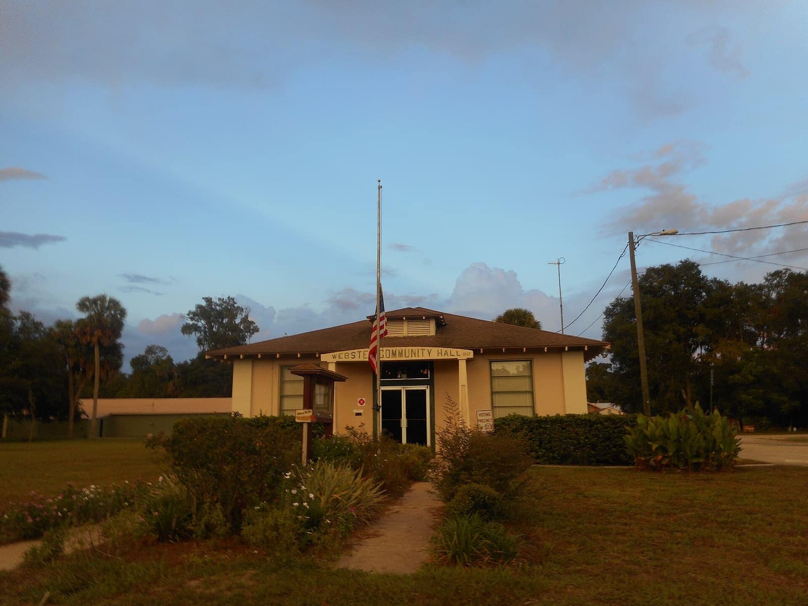 Single-storey community hall with an American flag at half-staff and shrubs at dusk.