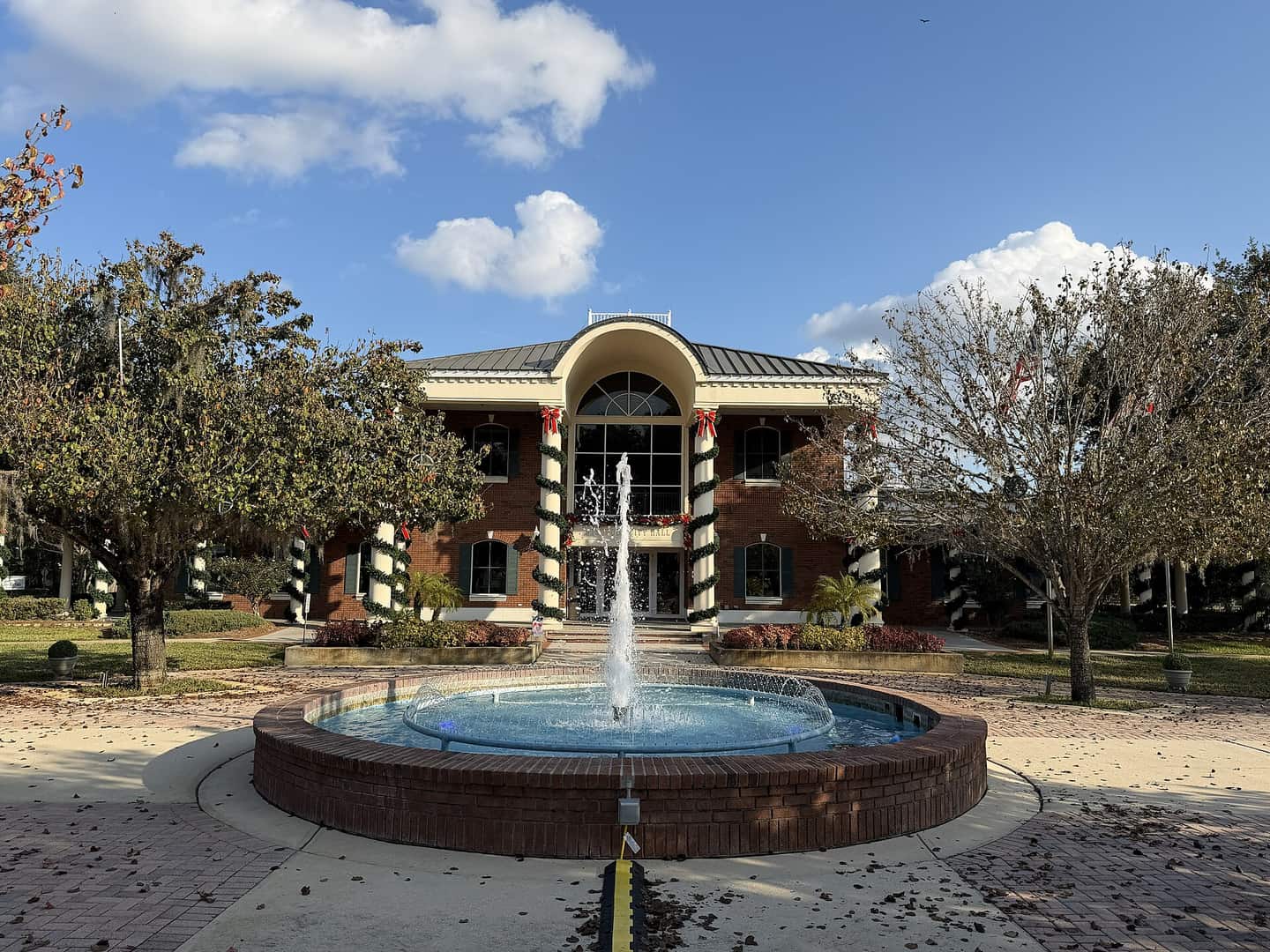Two-story brick government building with white columns and a round fountain in front, under blue sky.